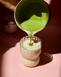 Matcha with perfect foaming being poured from a chawan into a glass on a pink background
